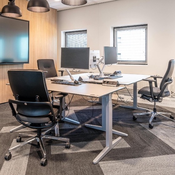 Modern office workspace with sit-stand desks, dual monitors, ergonomic seating, and wall display for collaborative pre-production.